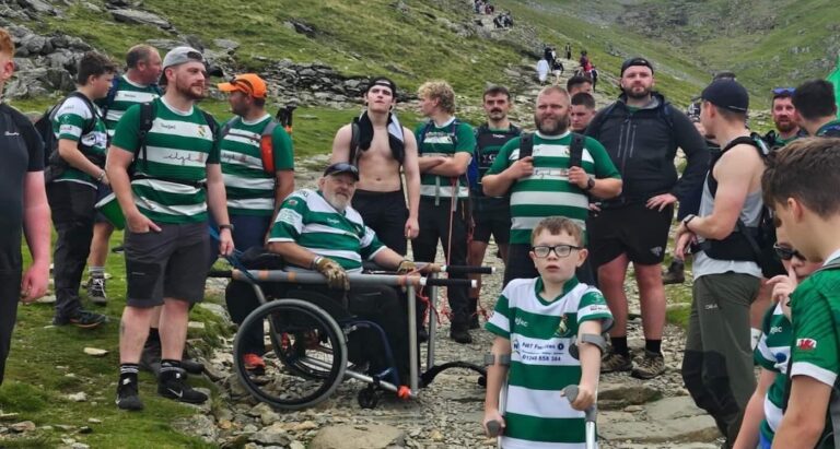 Members of a rugby team carrying a wheelchair-bound man on a rocky path up a mountain, with a scenic view in the background. They are working together with focus and determination.